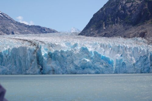 The end of a waterway blocked by the bulk of a glacier. The ice mass spans the width of the screen and beyond in white-blue snow pack, and sort-stained ice with a large dark crack slicing into the whole near the left hand side.