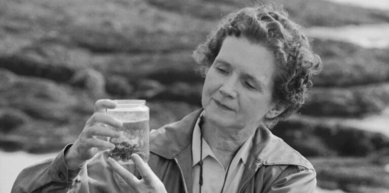 Black and white photo of a white woman with short, wavy hair on a rocky beach. She holds a jar filled with water and rocks.