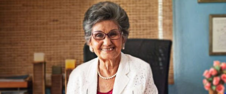 Professor Bonnelly, an older woman with light brown skin and salt-n-pepper hair. She wears a white blazer, red blouse, a strand of pearls, and glasses. She is smiling at her desk, books and papers before her. Photographer unkown.