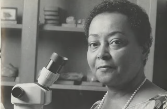Black and white photo of a Black woman with short, curly, dark hair seated at a microscope in a lab. She wears a floral blouse and a long sting of pearls. In her right hand she holds a button coral: a circle of white with faint striation radiating from the center