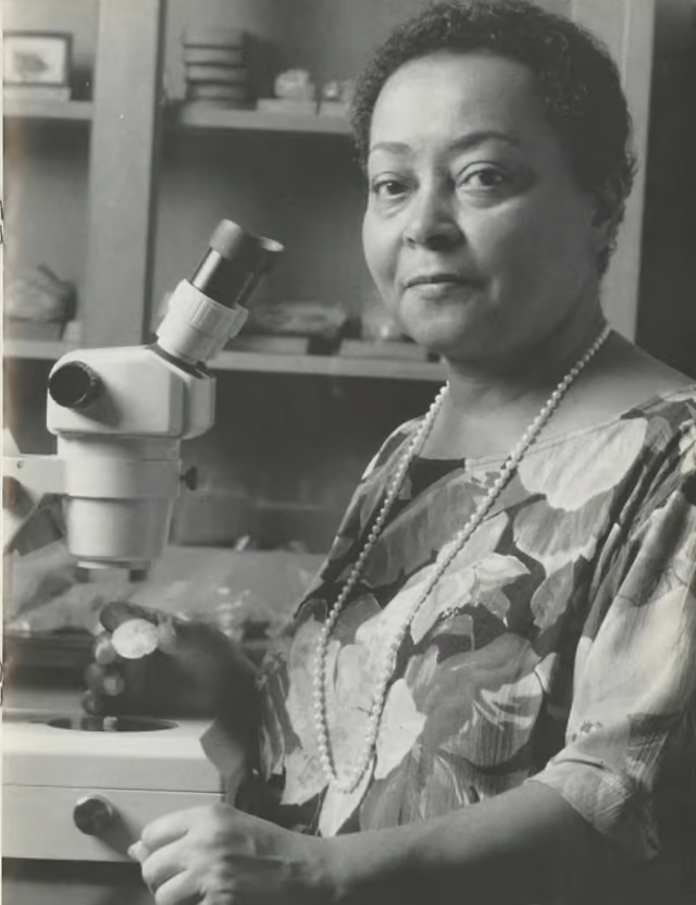 Black and white photo of a Black woman with short, curly, dark hair seated at a microscope in a lab. She wears a floral blouse and a long sting of pearls. In her right hand she holds a button coral: a circle of white with faint striation radiating from the center