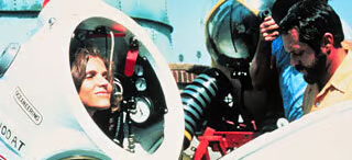 Dr. Sylvia Earle, a white woman with brown hair, stands inside a white, hard sided dive suite. Dials and tubes can be seen inside, near her head. Two men assist her from outside