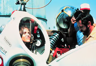  Dr. Sylvia Earle, a white woman with brown hair, stands inside a white, hard sided dive suite. Dials and tubes can be seen inside, near her head. Two men assist her from outside