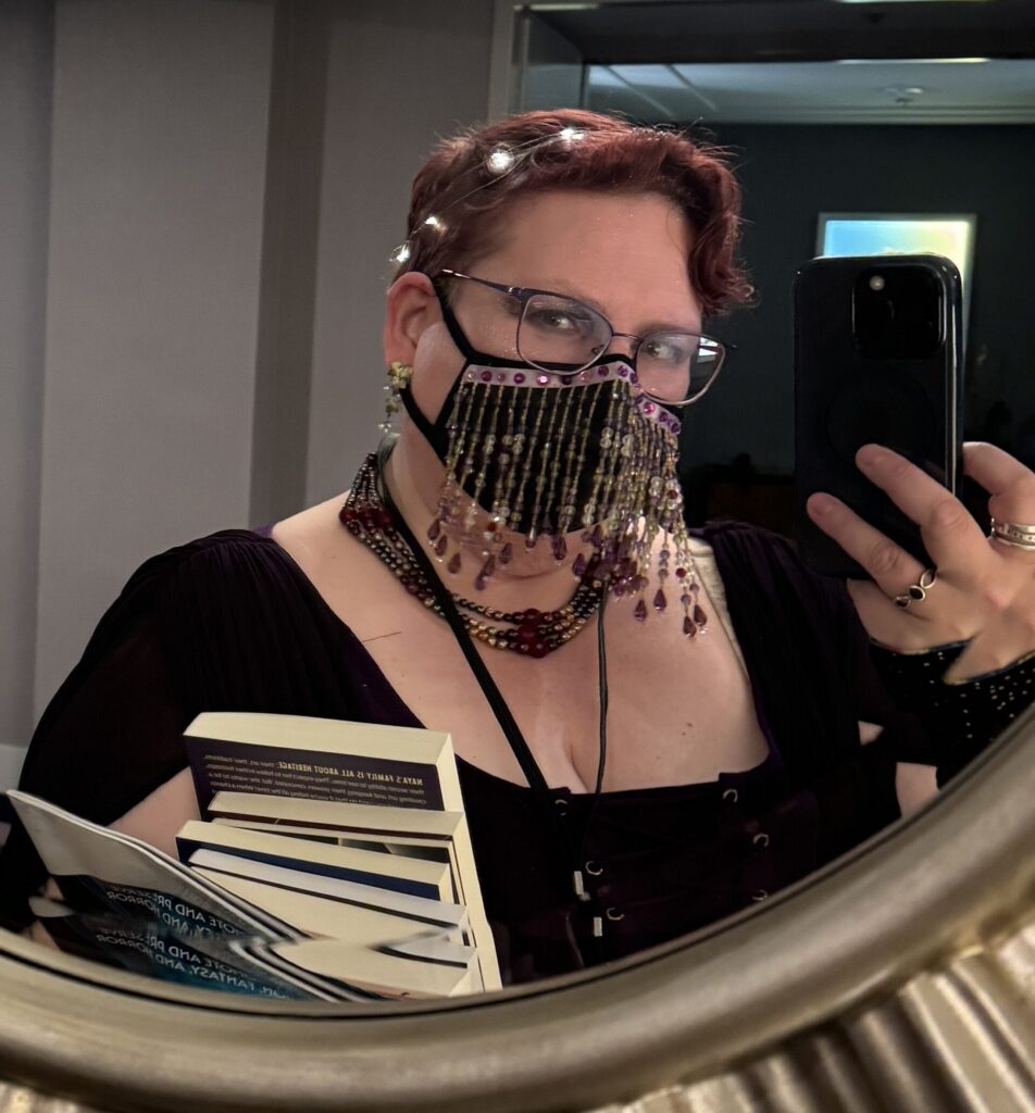 The author, a pale woman with short, dark cherry red hair with a wreath of fairy lights, is taking a selfie in a large round mirror. In one arm is a stack of books, in the other, her phone. She's wearing a dark purple dress, a chunky rope of dark red pearls at her neck, glasses, and a purple beaded facemask. 