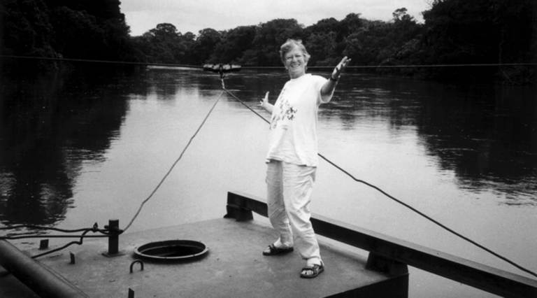 ID: Black and white photo of a smiling, middle aged, white woman with short hair, her arms out stretched. She’s wearing an over-sized white t-shirt, khaki pants and dark sandals. She stands on a flat surface (possibly a barge) on a smooth water way, surrounded by trees